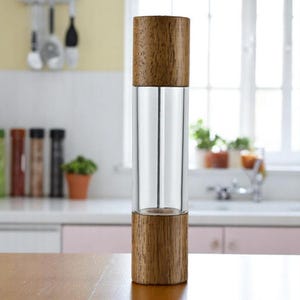 Op de afbeelding: Een pepermolen van helder glas en hout. De boven- en onderkant zijn gemaakt van lichtbruin hout, terwijl het midden een heldere glazen cilinder is. De molen staat op een houten oppervlak in een keuken.
