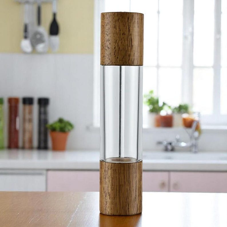 Op de afbeelding: Een pepermolen van helder glas en hout. De boven- en onderkant zijn gemaakt van lichtbruin hout, terwijl het midden een heldere glazen cilinder is. De molen staat op een houten oppervlak in een keuken.