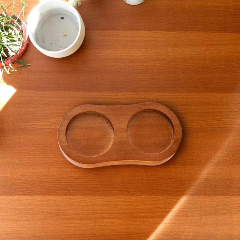 May include: A wooden coaster with two circular indentations, designed to hold two glasses or mugs. The coaster is made of a light brown wood and has a smooth, polished finish. The coaster is on a wooden surface.