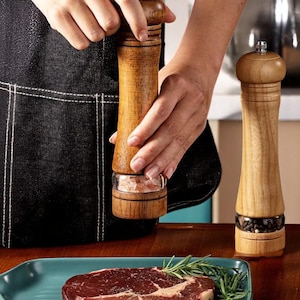 May include: Wooden salt and pepper grinders with a clear glass base. One grinder is being used over a teal plate with a raw steak and rosemary sprigs. The other grinder is standing upright. The grinders are made of light brown wood.