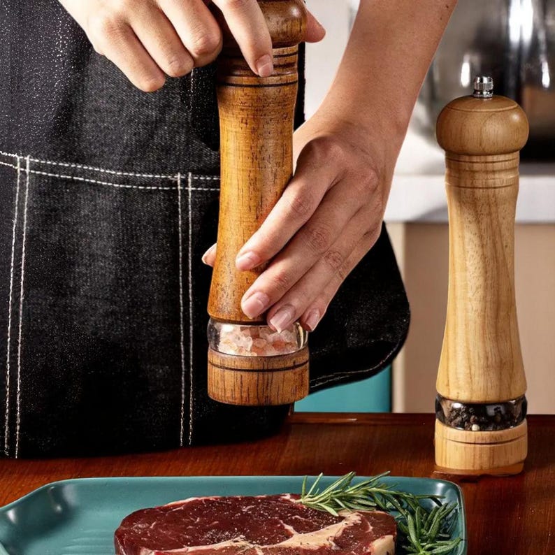 May include: Wooden salt and pepper grinders with a clear glass base. One grinder is being used over a teal plate with a raw steak and rosemary sprigs. The other grinder is standing upright. The grinders are made of light brown wood.