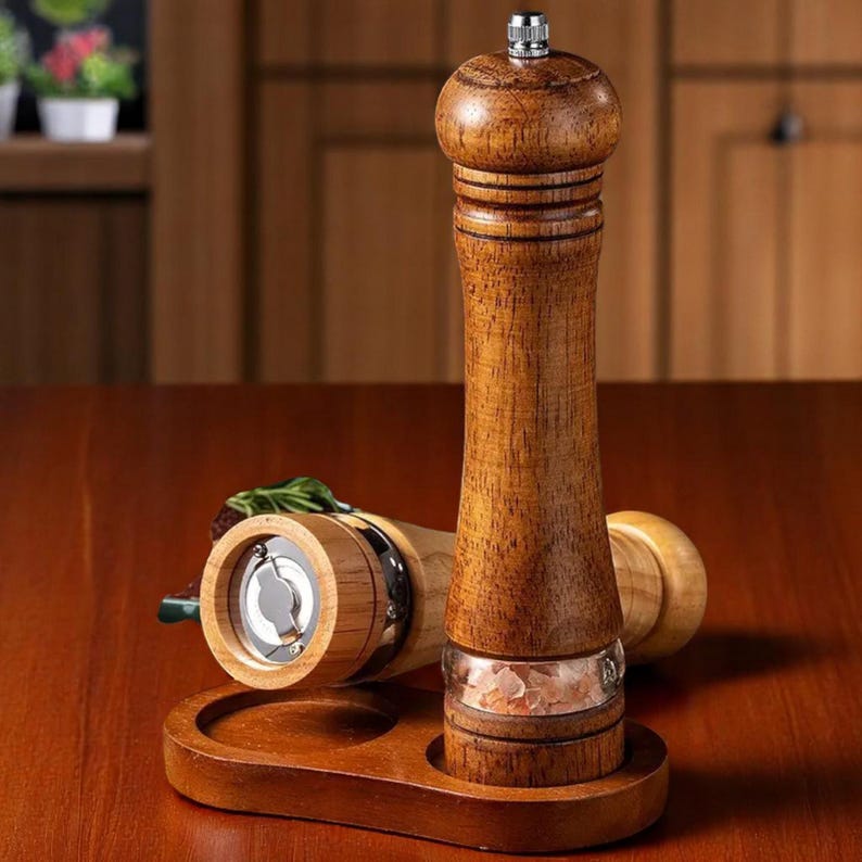 May include: A wooden salt and pepper grinder set. The grinders are made of light brown wood with a clear glass section to view the contents. The set includes a wooden base with two circular cutouts to hold the grinders. The grinders are on a dark brown table.