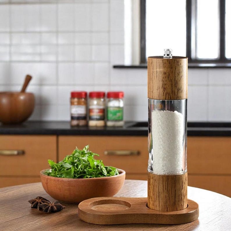 Op de afbeelding: Een houten zout- of pepermolen met een helder glazen midden gevuld met zout. De molen staat op een houten voet met een kleine cirkelvormige inkeping. Een houten kom met groene kruiden en een houten kom staan ook op de tafel.