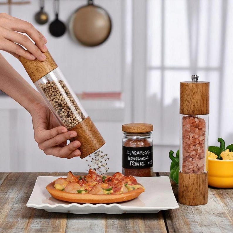 Op de afbeelding: Een houten en heldere acryl pepermolen die peperkorrels op een bord met eten doseert. Een glazen pot met een houten deksel en een zoutmolen zijn ook zichtbaar.