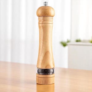 May include: A wooden pepper mill with a clear glass section showing black peppercorns. The mill has a light wood finish, a bulbous top, and a silver-colored metal cap. It stands on a wooden surface.