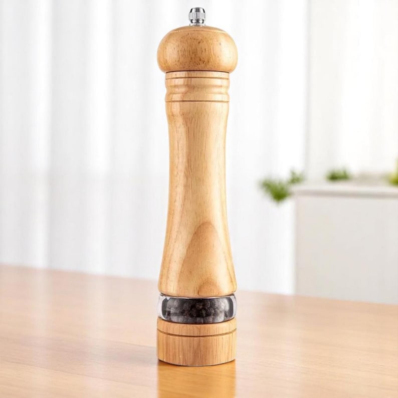 May include: A wooden pepper mill with a clear glass section showing black peppercorns. The mill has a light wood finish, a bulbous top, and a silver-colored metal cap. It stands on a wooden surface.