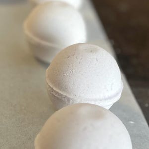 May include: A row of white, spherical bath bombs with a textured surface. The bath bombs are arranged in a line, with a soft, blurred background. The spherical shape is divided by a subtle line.