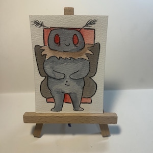 May include: A small watercolor painting on a wooden easel depicting a grey moth-like creature with large red eyes, a tan furry collar, and dark wings. The creature is centered against a rectangular red background on textured white paper.