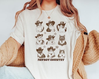 May include: A white t-shirt with a graphic of nine cats wearing cowboy hats and the text "Pawboy Country".