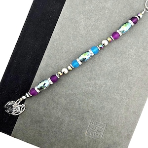 May include: A beaded bookmark with a silver charm. The bookmark features a series of beads in shades of purple, blue, and silver. The beads are strung together with a silver chain. The charm is a silver filigree design. The bookmark is resting on a gray surface.