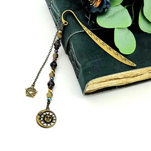 May include: A bronze bookmark with a crescent moon and sun charm, a pentagram charm, and a beaded chain. The bookmark is resting on a dark green book. The bookmark's hook has a decorative design.
