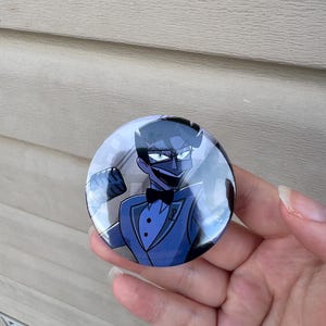 May include: A round button featuring a cartoon illustration of a character in a blue tuxedo with a black bow tie. The character has dark hair and is wearing a mask. The button is held in a hand.