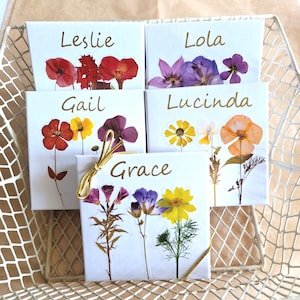 May include: Four white gift boxes with gold ribbon, each with a different name and pressed flower design. The names are Leslie, Lola, Gail, and Grace.