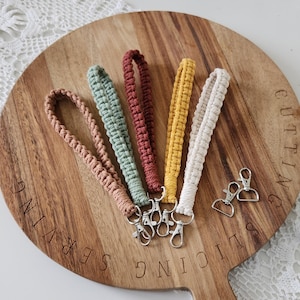 Puede incluir: Cinco llaveros de muñeca de macramé en tonos beige, verde, rojo, amarillo y blanco, cada uno con un cierre plateado. Los llaveros están colocados sobre una tabla de cortar de madera con texto grabado.