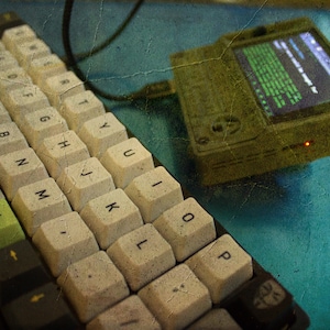 Peut inclure: Gros plan sur un clavier mécanique blanc avec des touches vertes et un petit appareil portable vert avec un écran affichant du texte. Le clavier et l'appareil sont sur une surface bleue.