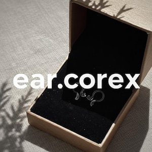 Puede incluir: Una pequeña caja de cartón abierta con un interior de terciopelo negro contiene un par de pendientes transparentes y curvados. Los pendientes tienen forma de serpiente. El texto "ear.corex" está impreso en blanco en la caja.