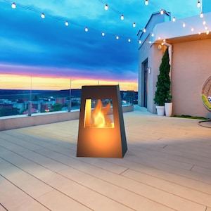 Könnte beinhalten: Ein moderner Außenkamin mit brennender Flamme auf einer Holzterrasse. Die Szene wird von Lichterketten vor einem Abendhimmel beleuchtet. Ein Hängesessel und Topfpflanzen sind ebenfalls zu sehen.
