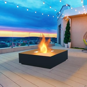 Puede incluir: Escena de patio exterior moderno al anochecer. Una hoguera cuadrada negra con una llama brillante se encuentra en una terraza de madera. Luces de cadena iluminan el espacio. Una silla colgante y plantas en macetas contribuyen al ambiente.