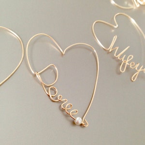 May include: Two gold wire heart-shaped ornaments with the names "Wifey" and "Darrel" spelled out in wire. The "Darrel" ornament has a small pearl at the bottom.