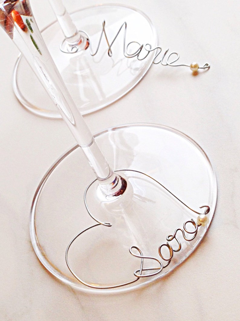 May include: Two clear glass wine glasses with wire name tags. The name "Marie" is on one glass and the name "Sara" is on the other glass.