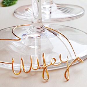 May include: A gold wire word "Wife" is wrapped around the stem of a clear wine glass.