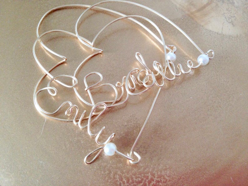 May include: Gold wire heart-shaped ornament with the names "Emily" and "Bradley" spelled out in wire. The ornament is decorated with small white pearls.