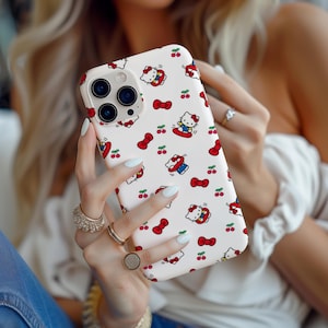 Może przedstawiać: Białe etui na iPhone'a z powtarzającym się wzorem postaci Hello Kitty, czerwonych kokardek i wiśni. Etui ma błyszczące wykończenie i jest trzymane w dłoniach osoby. Etui ma wycięcie na aparat.