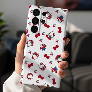 Może przedstawiać: Białe etui na telefon z powtarzającym się wzorem postaci z kresk&oacute;wki kota, czerwonych kokardek i wiśni. Etui na telefon ma system potr&oacute;jnego obiektywu aparatu. Projekt jest zabawny i kolorowy.