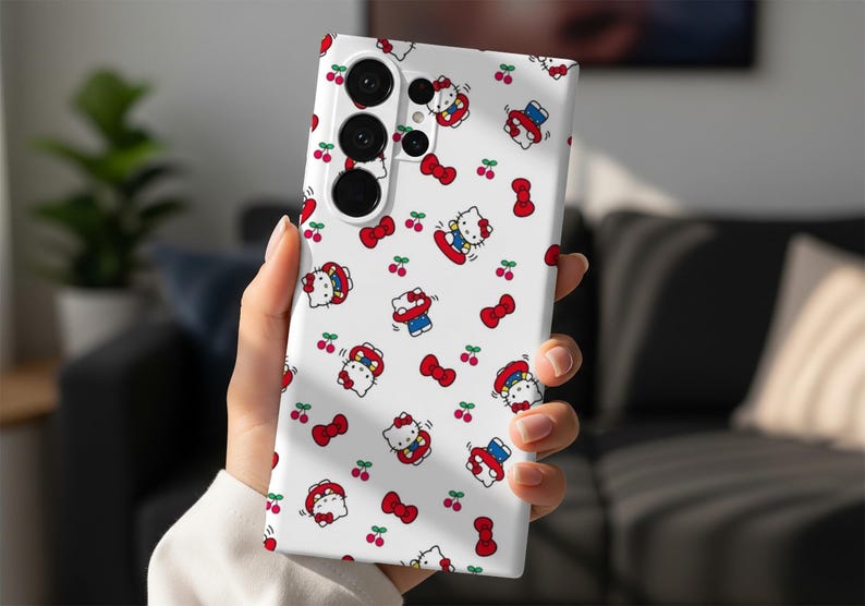 Może przedstawiać: Białe etui na telefon z powtarzającym się wzorem postaci z kresk&oacute;wki kota, czerwonych kokardek i wiśni. Etui na telefon ma system potr&oacute;jnego obiektywu aparatu. Projekt jest zabawny i kolorowy.