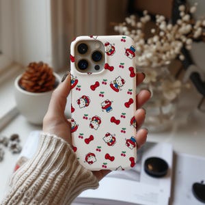 Może przedstawiać: Białe etui na telefon z powtarzającym się wzorem postaci z kresk&oacute;wki kota, czerwonych wiśni i czerwonych kokardek. Etui ma wycięcie na aparat i jest trzymane w dłoni osoby.