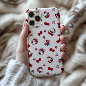 Może przedstawiać: Białe etui na iPhone'a z powtarzającym się wzorem postaci Hello Kitty, czerwonych kokardek i wiśni. Etui ma błyszczące wykończenie i wycięcie na aparat.