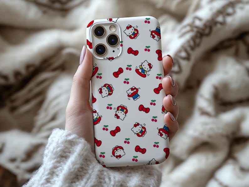 Może przedstawiać: Białe etui na iPhone'a z powtarzającym się wzorem postaci Hello Kitty, czerwonych kokardek i wiśni. Etui ma błyszczące wykończenie i wycięcie na aparat.