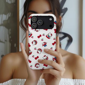 Może przedstawiać: Białe etui na telefon z powtarzającym się wzorem postaci Hello Kitty, czerwonych kokardek i wiśni. Etui ma czarny moduł aparatu. Etui na telefon jest trzymane przed osobą.