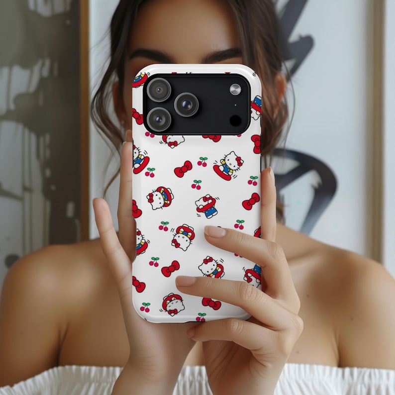 Może przedstawiać: Białe etui na telefon z powtarzającym się wzorem postaci Hello Kitty, czerwonych kokardek i wiśni. Etui ma czarny moduł aparatu. Etui na telefon jest trzymane przed osobą.