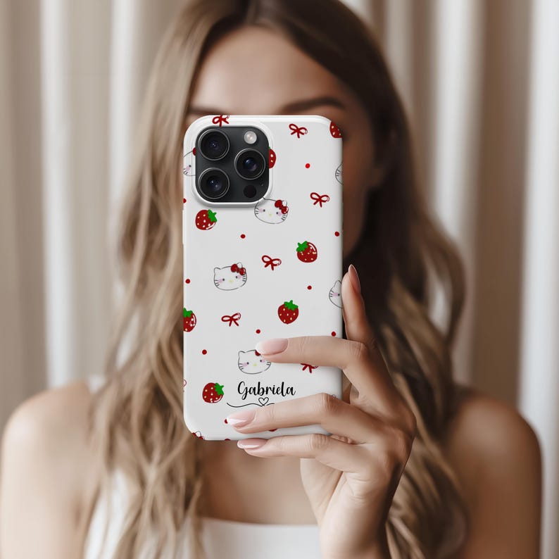 May include: White phone case featuring a pattern of red strawberries, red bows, and cartoon cat faces. The name "Gabriela" is written in black script with a small heart below it. The case has a camera cutout.