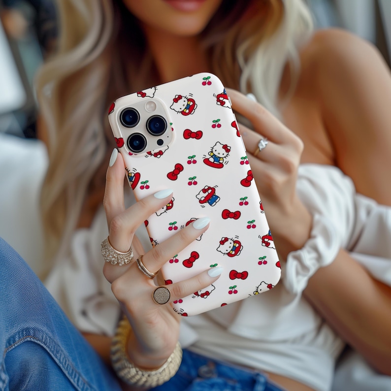 Może przedstawiać: Białe etui na iPhone'a z powtarzającym się wzorem postaci Hello Kitty, czerwonych kokardek i wiśni. Etui ma błyszczące wykończenie i jest trzymane w dłoniach osoby. Etui ma wycięcie na aparat.