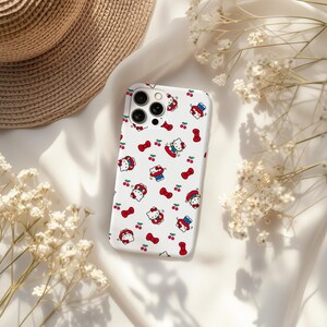 Może przedstawiać: Białe etui na telefon z powtarzającym się wzorem postaci Hello Kitty, czerwonych wiśni i czerwonych kokardek. Etui ma wycięcie na aparat. Białe kwiaty i słomkowy kapelusz w tle.