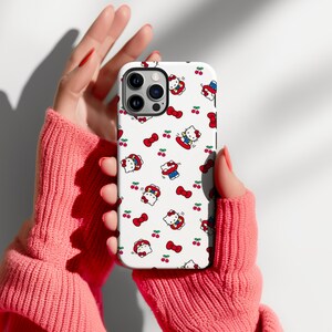 Może przedstawiać: Białe etui na telefon z powtarzającym się wzorem postaci Hello Kitty, czerwonych kokardek i wiśni. Etui ma błyszczące wykończenie i jest przeznaczone do smartfona. Projekt zawiera Hello Kitty w r&oacute;żnych strojach i pozach.