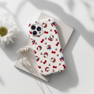 Może przedstawiać: Białe etui na telefon z powtarzającym się wzorem postaci z kresk&oacute;wki kota, czerwonych kokardek i wiśni. Etui ma wycięcie na aparat i spoczywa na białej powierzchni, z kwiatami w tle.