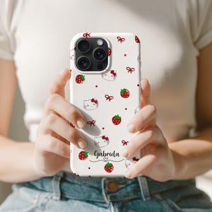 May include: White phone case featuring a pattern of red strawberries, bows, and cartoon cat faces. The name "Gabriela" is written in a script font. The case has a glossy finish and is designed for a smartphone.
