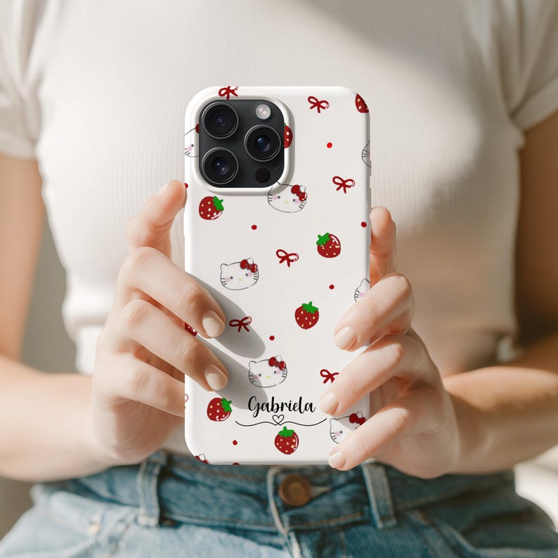 May include: White phone case featuring a pattern of red strawberries, bows, and cartoon cat faces. The name "Gabriela" is written in a script font. The case has a glossy finish and is designed for a smartphone.