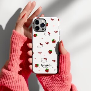 May include: A white phone case featuring a pattern of red strawberries, bows, and cartoon cat faces. The name "Gabriela" is written in cursive with a heart below. The case is held in a person's hands.