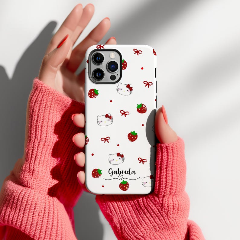 May include: A white phone case featuring a pattern of red strawberries, bows, and cartoon cat faces. The name "Gabriela" is written in cursive with a heart below. The case is held in a person's hands.