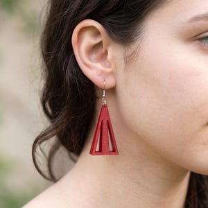 May include: A red earring featuring a geometric triangular frame with a central vertical bar, suspended from a silver-toned metal hook. The earring has a matte finish and a hollow design, creating a distinct shape that hangs from the earlobe.