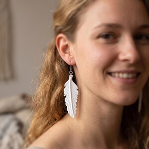 Può includere: Un paio di orecchini a forma di piuma bianca con una superficie dettagliata e strutturata che imita l'aspetto di un piumaggio naturale. Ogni orecchino pende da un semplice gancio in metallo color argento, per un look pulito e distinto.