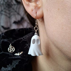Op de afbeelding: Een witte spookvormige oorbel met een gladde, matte afwerking en twee ovale uitsparingen voor de ogen. De spookbedel hangt aan een zilverkleurige metalen haak en een kleine ronde ring, wat zorgt voor een hangend ontwerp voor de oorlel.