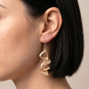 May include: A drop earring featuring a series of three interconnected, textured, light beige triangular shapes suspended from a silver-toned metal hook. The earring has a matte finish and a layered, geometric design that hangs vertically from the earlobe.