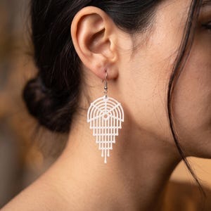 Può includere: Un orecchino pendente bianco dal design geometrico a ventaglio, caratterizzato da linee arcuate concentriche e barre verticali che si restringono verso il basso. L'orecchino è fissato a un gancio in metallo color argento.
