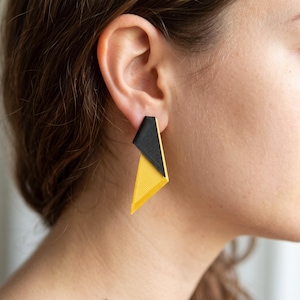 May include: A geometric drop earring featuring a two-tone design with a matte black upper triangular section and a textured bright yellow lower triangular section. The earring has a sharp, angular silhouette with clean lines and a flat surface.