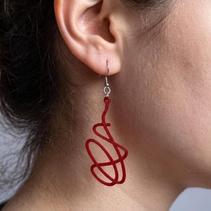 Peut inclure: Une boucle d'oreille pendante composée d'une forme abstraite rouge foncé, texturée, qui s'entrelace en courbes irrégulières et ajourées. L'élément est suspendu à un crochet métallique de couleur argentée qui traverse le lobe de l'oreille.